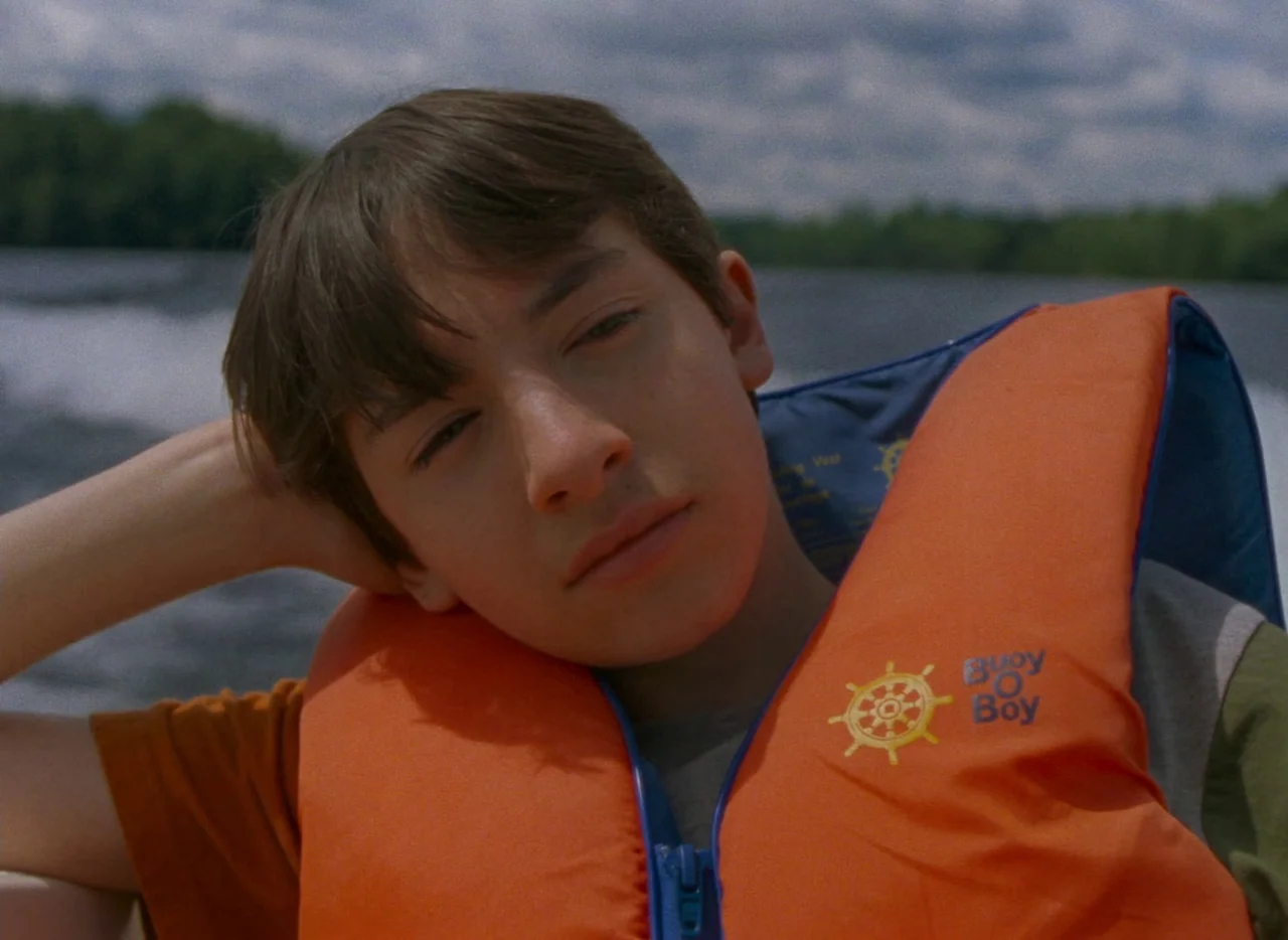 Falcon Lake (2022) movie still: closeup — A person sits on a boat with water and trees visible in the background. The individual wears an orange…
