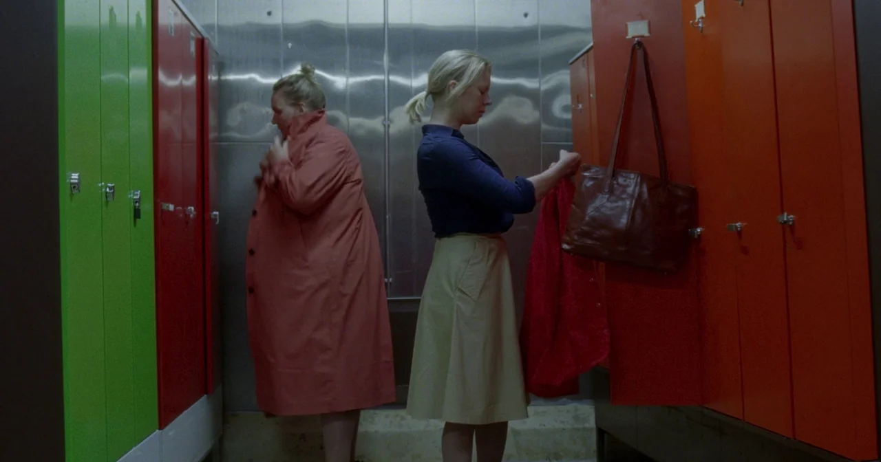 Fallen Leaves (2023) movie still: fullbody — A locker room contains rows of bright red and green lockers against stainless steel panels. Two women…