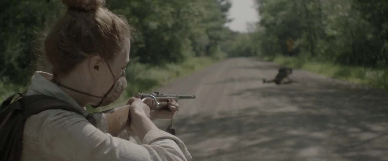 Here Alone movie still: medium — A rural, unpaved gravel road passes through dense deciduous trees on a summer day. A woman wearing a backpack and…