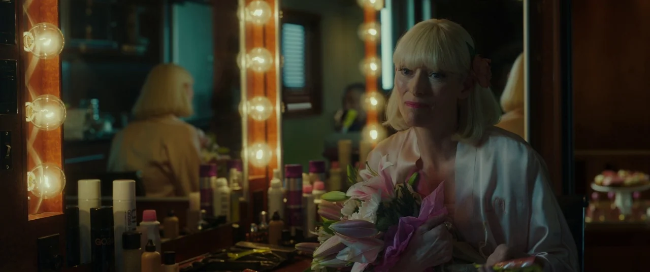 Okja (2017) movie still: medium — A dimly lit dressing room features a vanity mirror framed by incandescent bulbs. A woman in a pale silk robe holds…