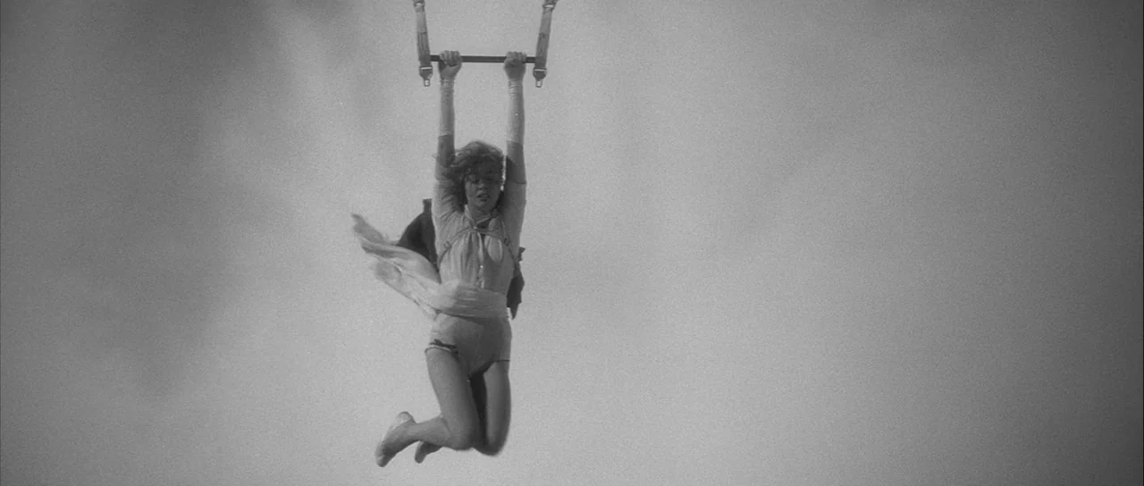 The Tarnished Angels (1957) movie still: fullbody — A trapeze artist swings through the air against a bright, plain sky backdrop. The woman holds…