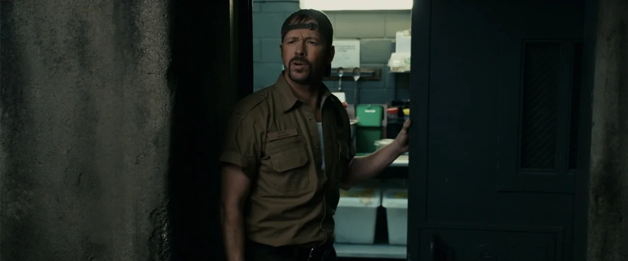 Zookeeper (2011) movie still: medium — A dimly lit room with concrete block walls features a man standing in a doorway. A man wearing a uniform and…