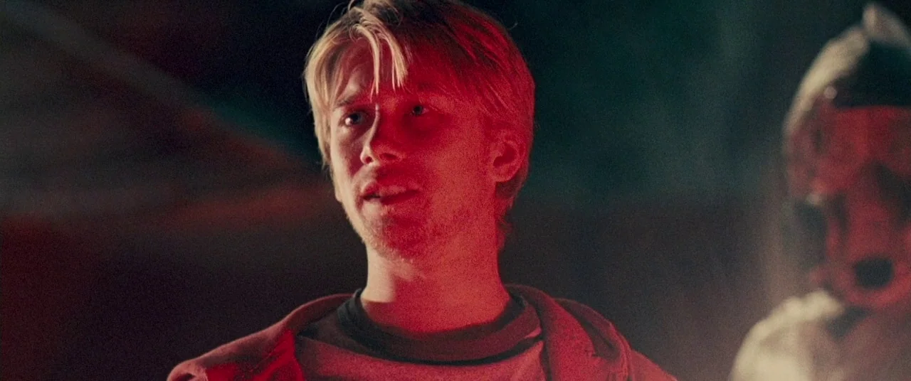 Carriers (2009) movie still: medium — A dark night setting lit by a bright, directional red light source. A young blond man stands in the light…