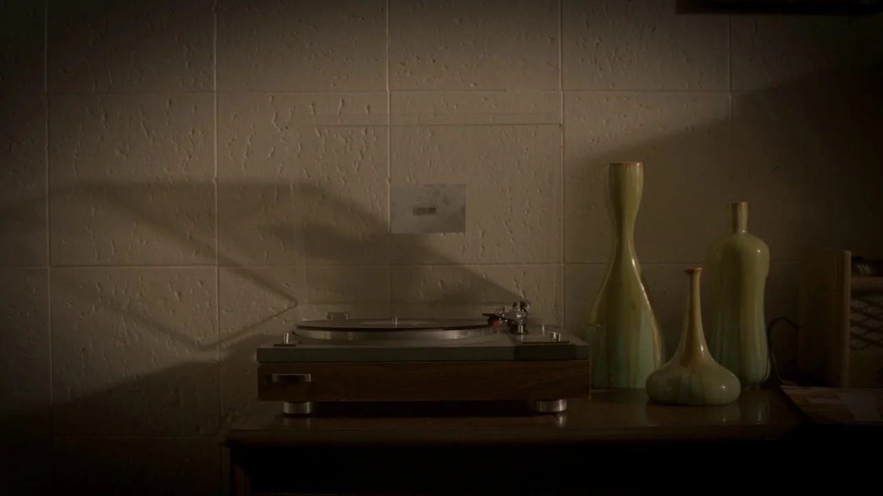 The Pact II movie still: medium — A vintage turntable and three ceramic vases are arranged on a dark wooden surface. A textured white wall forms the…