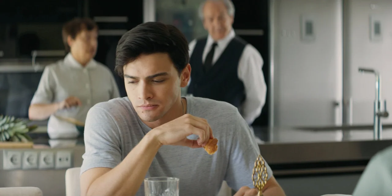 My Fault (2023) movie still: medium — A modern white kitchen features a young man in the foreground at a table. An elderly man in a suit and a woman…