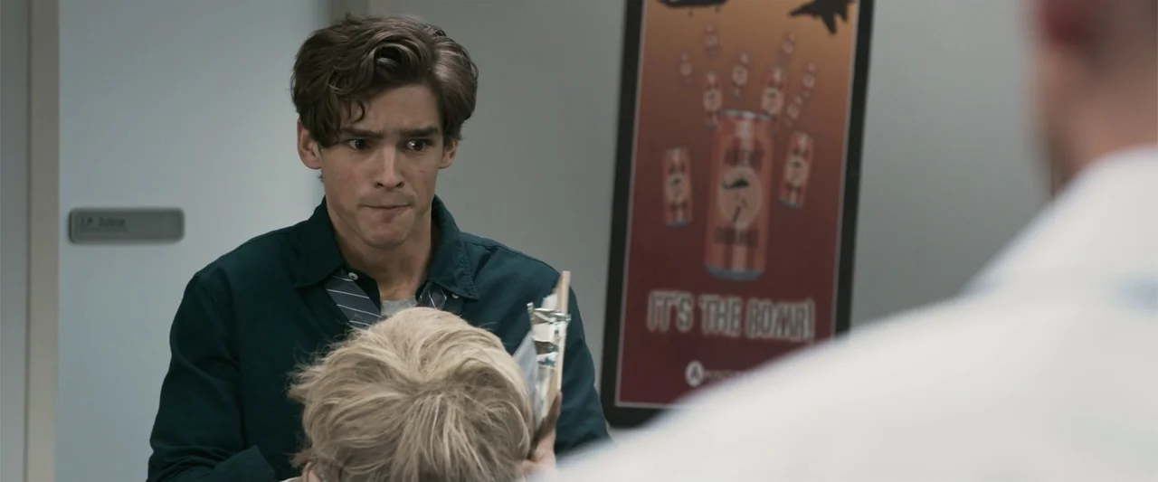 Office Uprising (2018) movie still: medium — An interior hallway features a white door with a name tag and a poster. A young man wearing a dark teal…