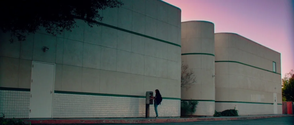 White Bird in a Blizzard (2014) movie still: wide — A modern commercial building with smooth panels and tiled lower sections sits at dusk. A woman…