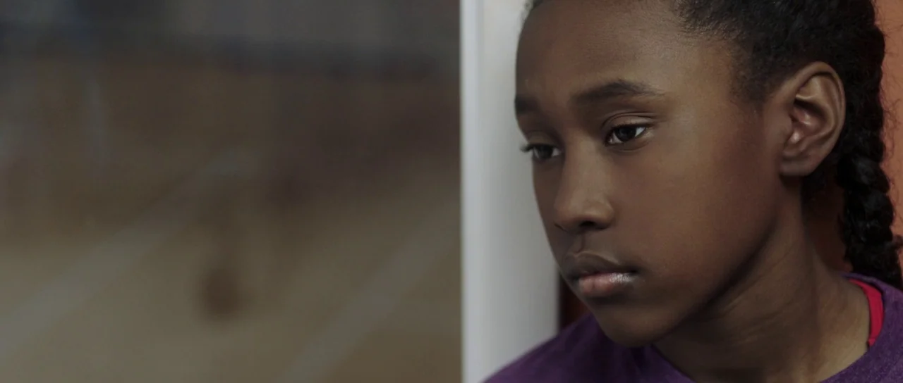The Fits (2016) movie still: closeup — A young girl sits by a window inside a transit carriage. She looks out the glass while the background appears…