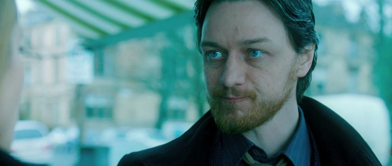 Filth (2013) movie still: medium — A public bus stop shelter sits under a green canopy with blurred street activity. A man with a ginger beard looks…