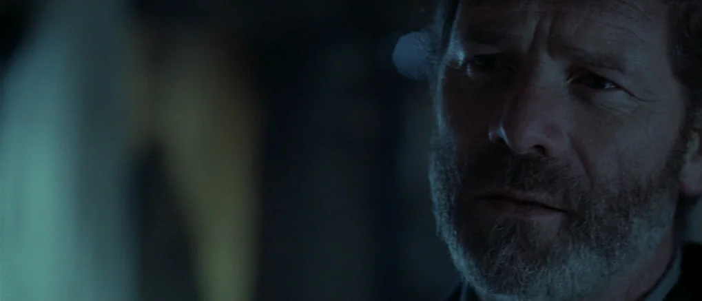 The Claim (2000) movie still: closeup — A dimly lit, indistinct interior space is shrouded in deep, cool-toned shadows. A middle-aged man with a…