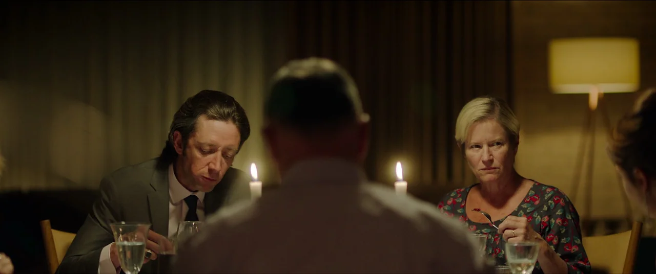 The Feast (2021) movie still: medium — A dimly lit room features three individuals seated around a table for dinner. They are eating silently while…