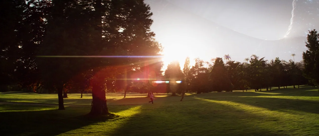 Elysium (2013) movie still: wide — A sprawling park with lush green grass and several mature evergreen trees. A large artificial celestial ring…