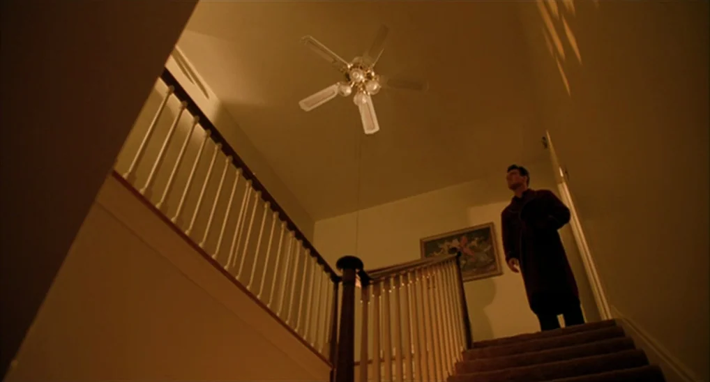 Twin Peaks: Fire Walk With Me (1992) movie still: wide — A dark, warm-toned staircase in a house featuring a central ceiling fan. A man stands on…