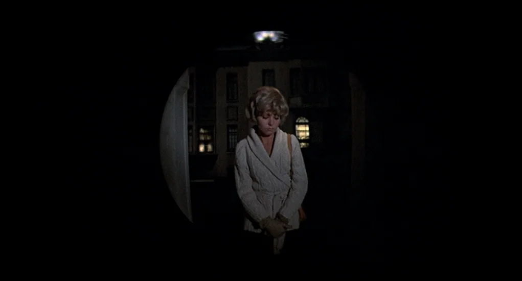 Family Plot (1976) movie still: medium — A woman stands before the dark doorway of an apartment building at night. The view is framed by a circular…