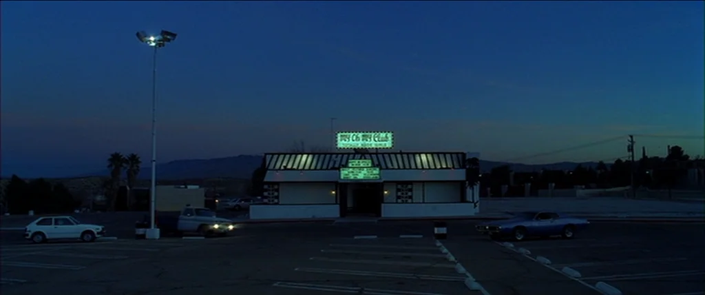 Kill Bill Vol. 2 (2004) movie still: establishing — An empty parking lot sits in front of a low-profile building with a neon sign. Several parked…