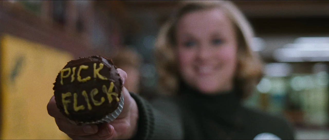 Election (1999) movie still: closeup — A woman holds out a chocolate frosted cupcake with yellow text in an aisle. The foreground object is in sharp…