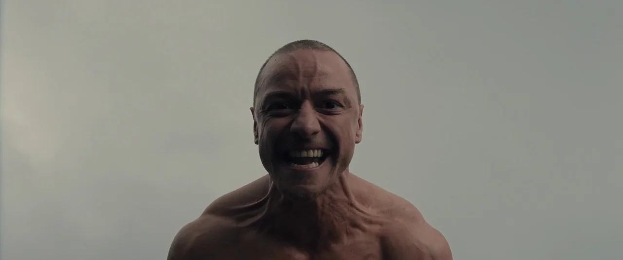 Glass (2019) movie still: closeup — A man stands against a plain, overcast sky with no visible horizon line. The subject faces the camera directly…