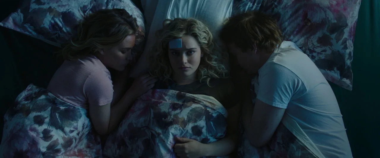 Spontaneous (2020) movie still: medium — A bedroom contains three people lying closely together in a bed under floral bedding. The woman in the…
