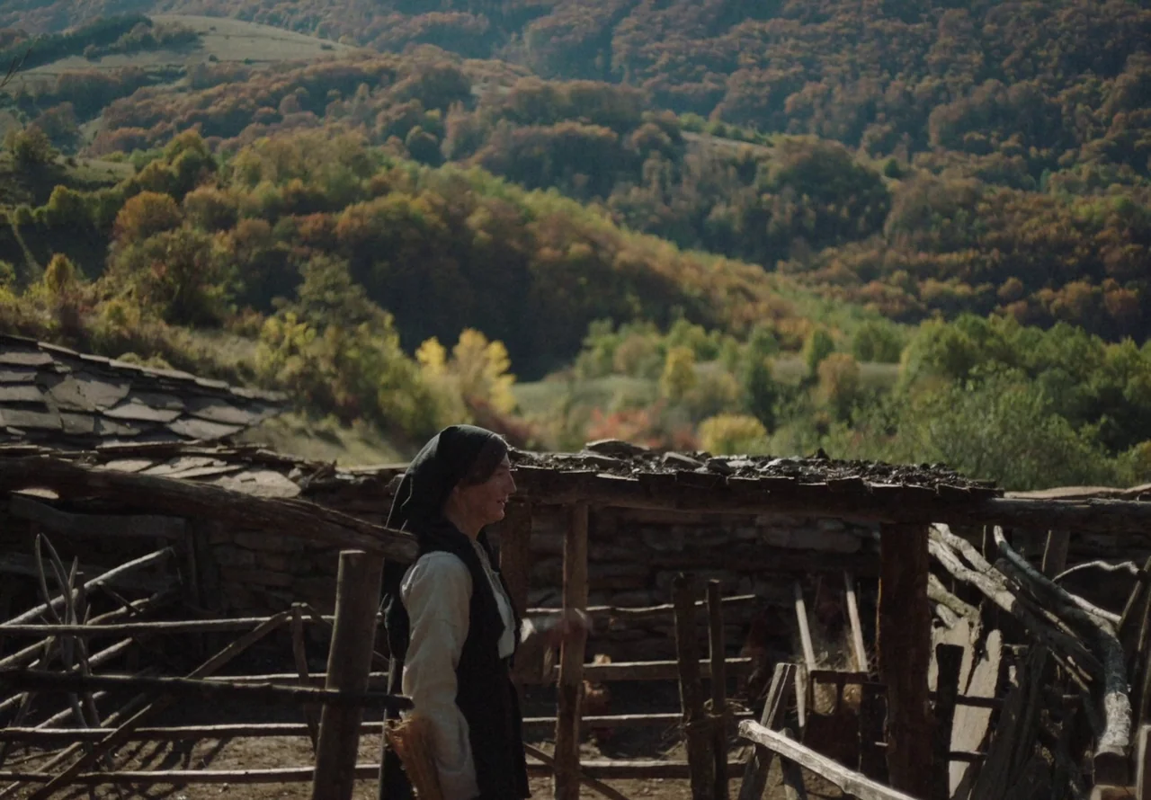 You Won’t Be Alone (2022) movie still: medium — A rural farm enclosure features rustic wooden fencing and a weathered structure. A woman wearing a…