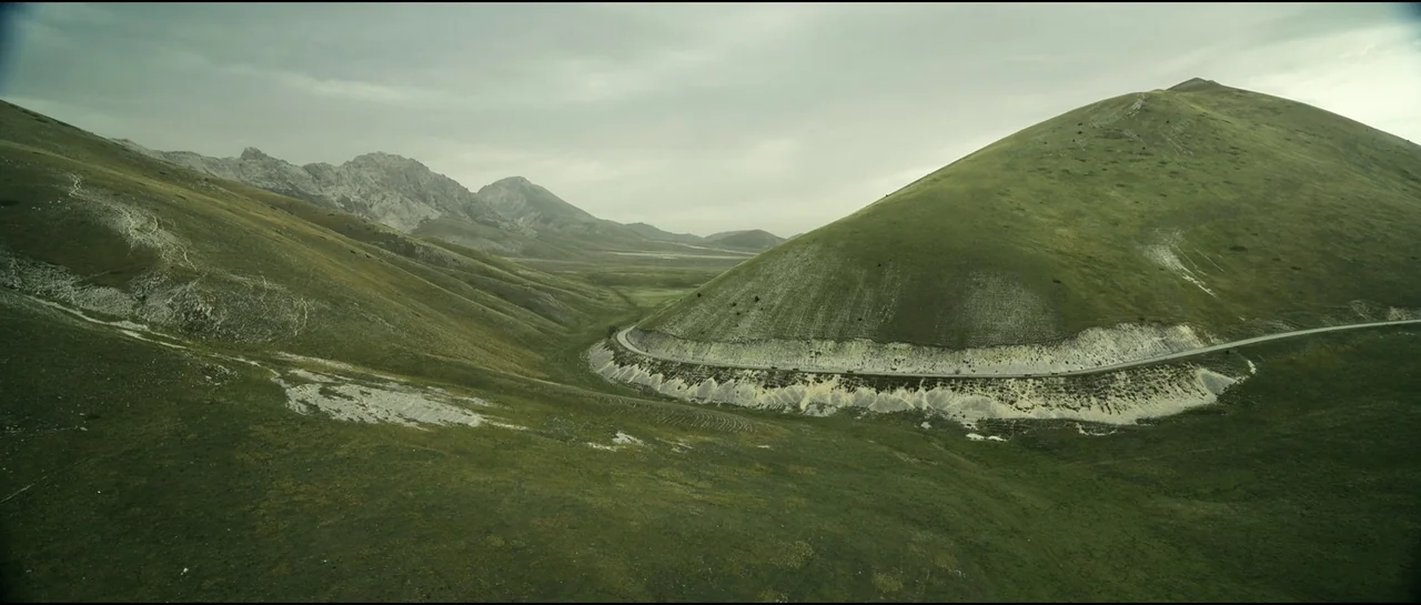 Ferrari (2023) movie still: establishing — A wide valley features rolling green hills with a paved road cutting across them. The background shows…