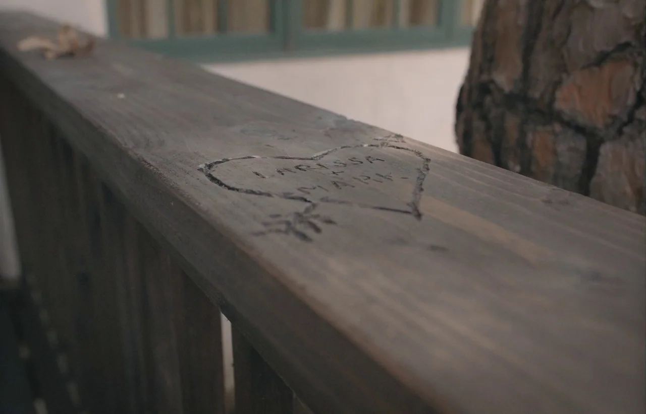 Marcel the Shell with Shoes On (2021) movie still: closeup — A rustic dark-stained wooden porch railing sits in front of a tree trunk. A heart shape…