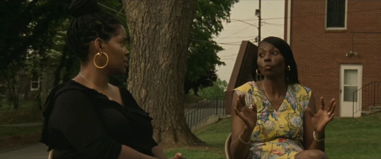 Sermon on the Mount (2019) movie still: medium — A church exterior with a large tree and brick building in the background. Two women sit in outdoor…