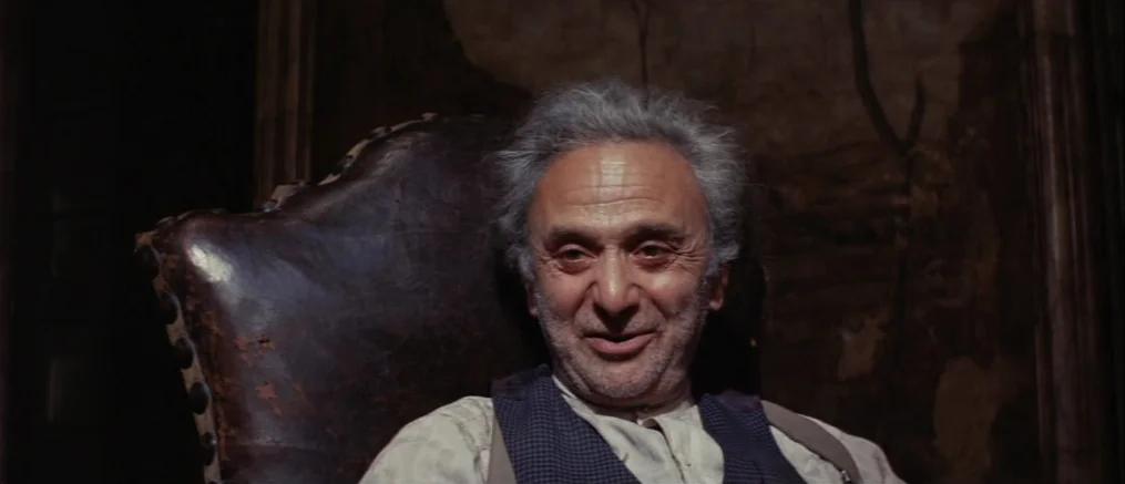 Catch 22 (1970) movie still: closeup — An older man sits in a worn leather chair in a dark, dimly lit room. The man has grey hair and wears a vest…