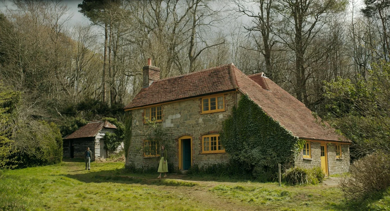 My Policeman (2022) movie still: wide — A stone house with a red tiled roof stands in a field. A woman walks near the entrance while a man stands by…
