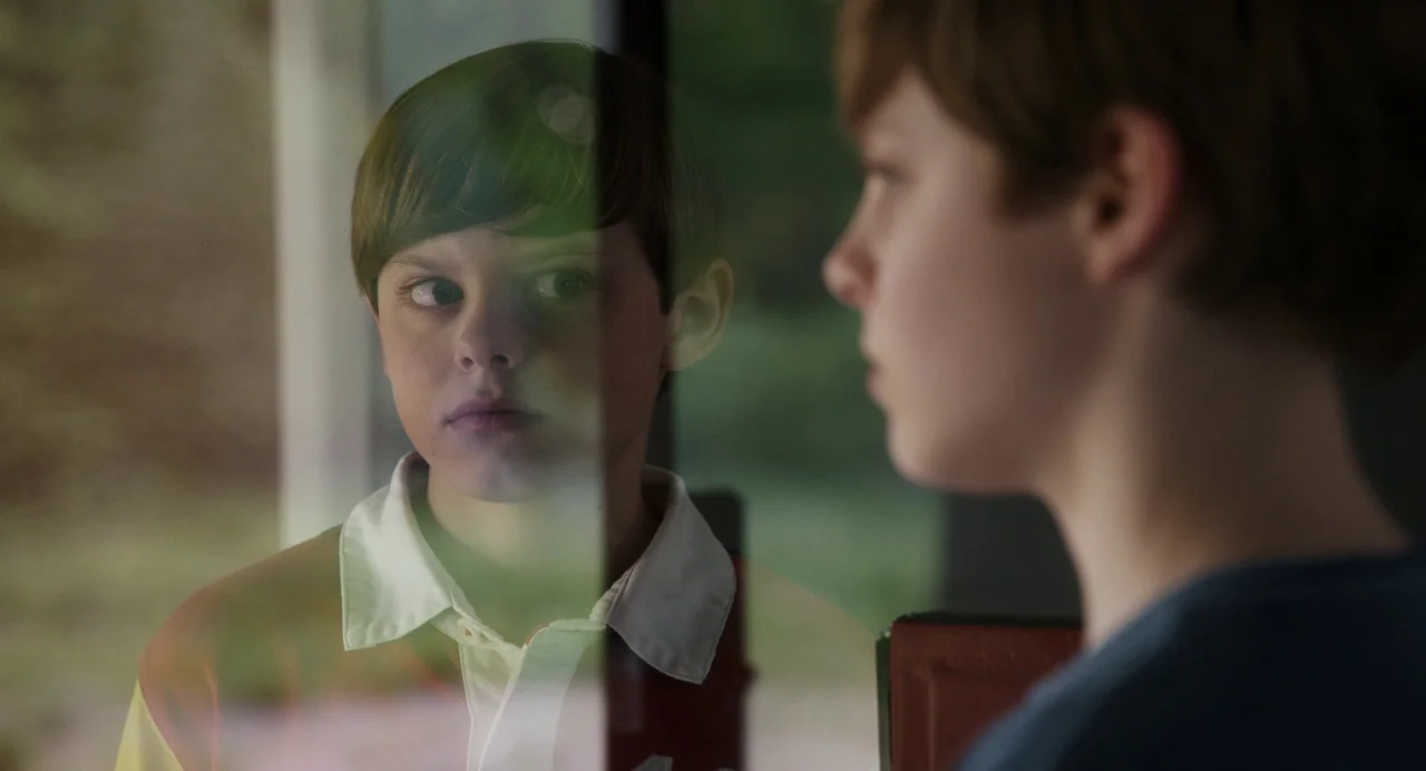 Goodnight Mommy (2022) movie still: medium — A young boy looks out through a large glass window pane during the daytime. The boy's face is mirrored…