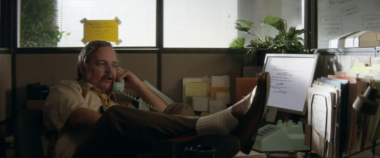 Seven Stages to Achieve Eternal Bliss (2018) movie still: medium — A cluttered 1970s cubicle is filled with paperwork, a green telephone, and office…