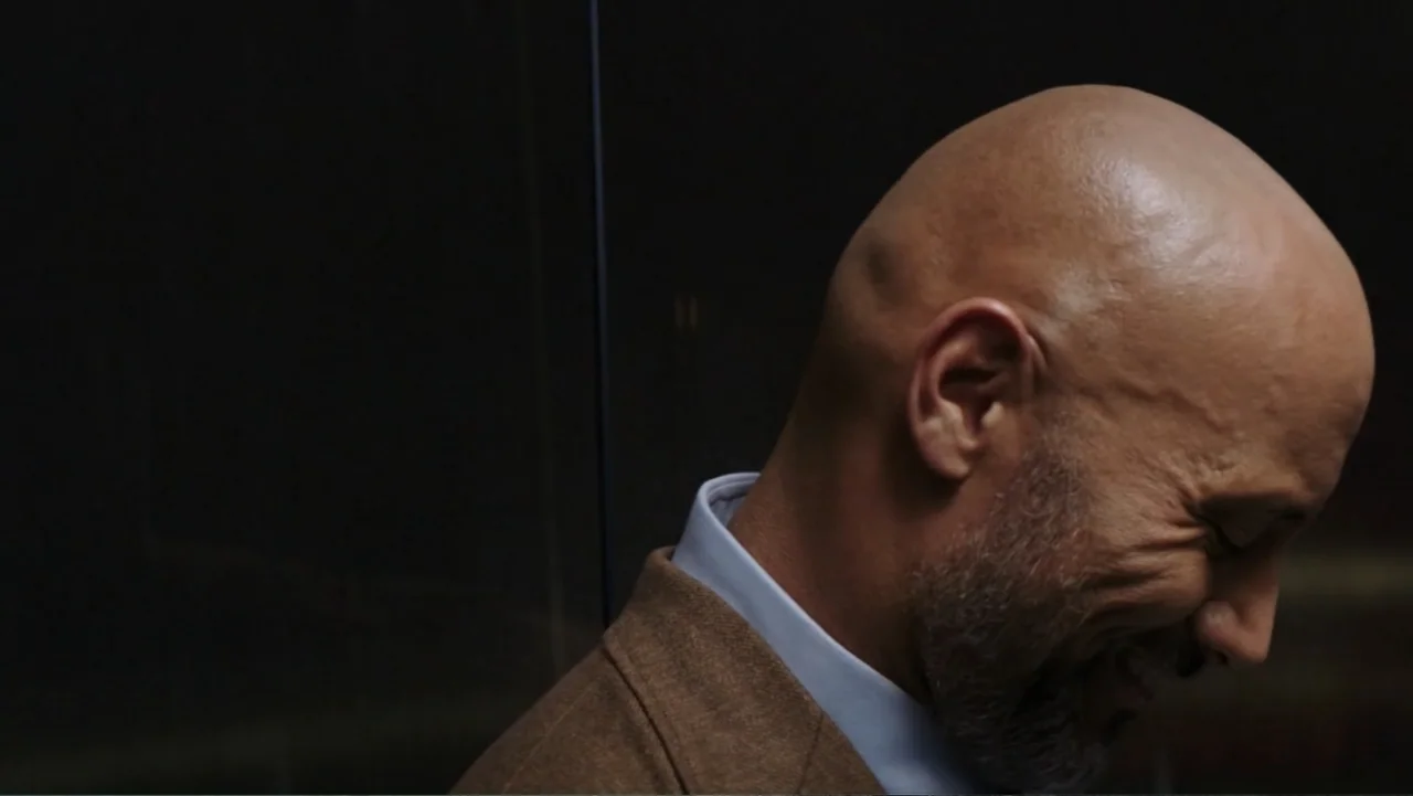 Skyscraper (2018) movie still: closeup — A dark interior setting with a smooth glass or dark wall surface visible behind. A bald man in a tan blazer…