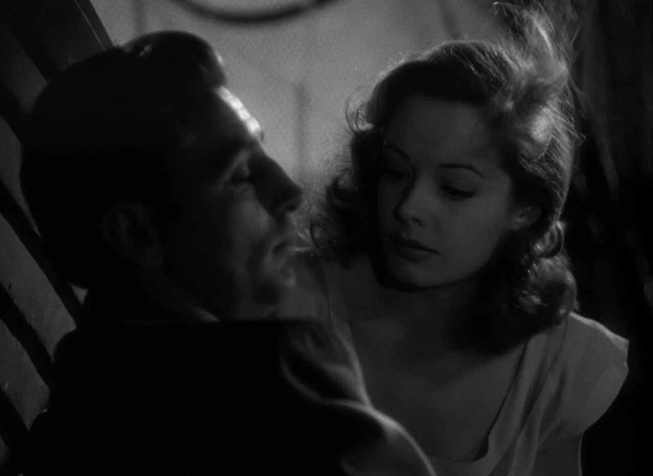 Out of the Past (1947) movie still: medium — A dark indoor space contains a man seated in a wooden chair. A woman sits nearby and leans in toward him.