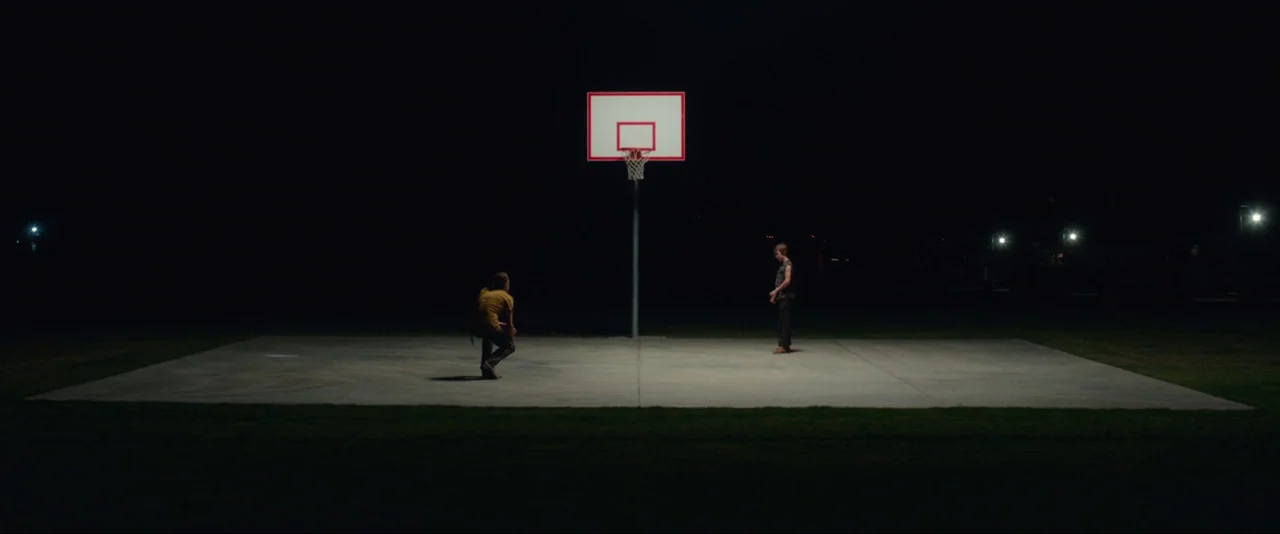 Captain Fantastic (2016) movie still: wide — A flat concrete court sits isolated in the darkness at night. Two people stand near a basketball hoop.
