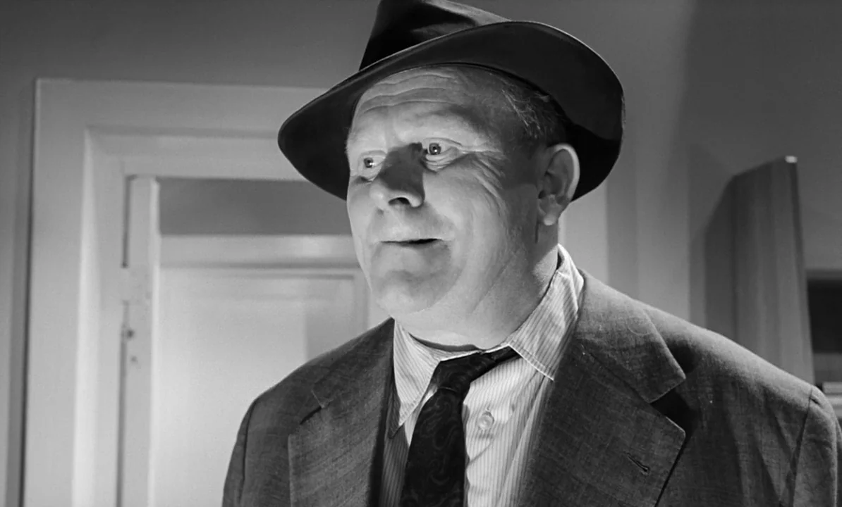 The 1,000 Eyes of Dr. Mabuse (1960) movie still: closeup — An interior space with a doorway visible in the blurred background. A middle-aged man…