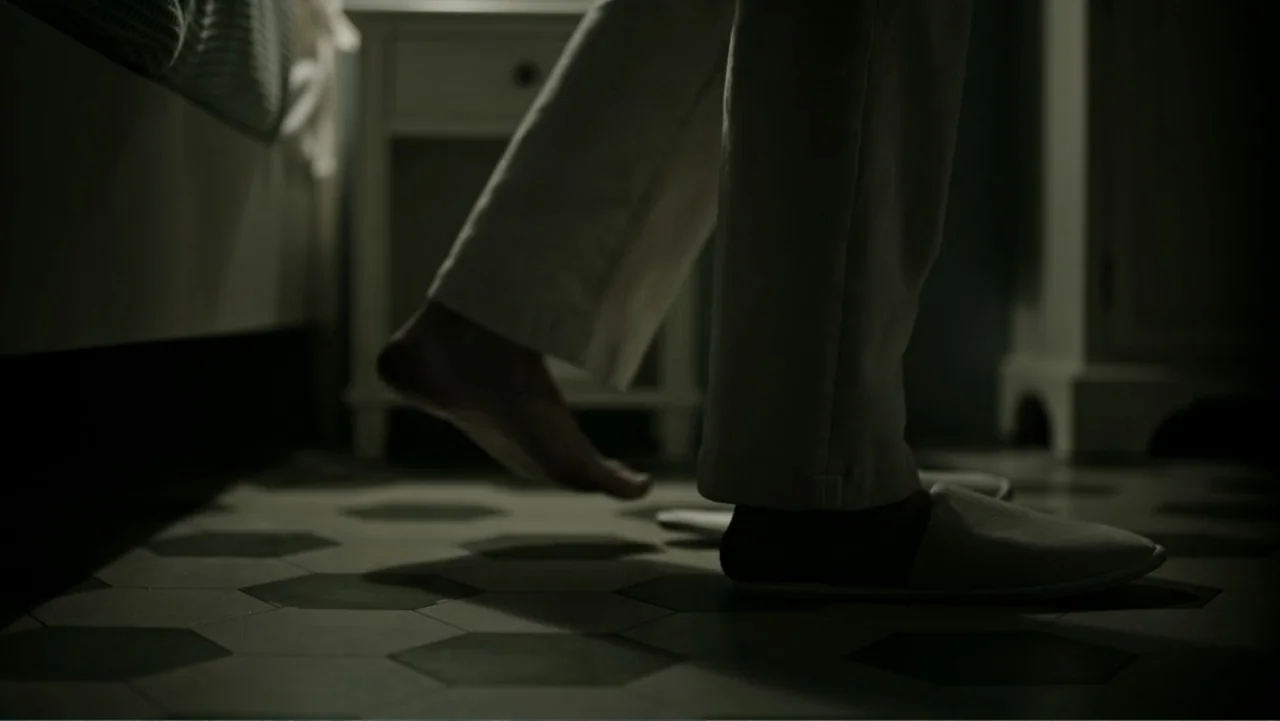 A Cure for Wellness (2017) movie still: closeup — A bedroom interior features dark floor tiles and white bedside furniture. A person wearing light…