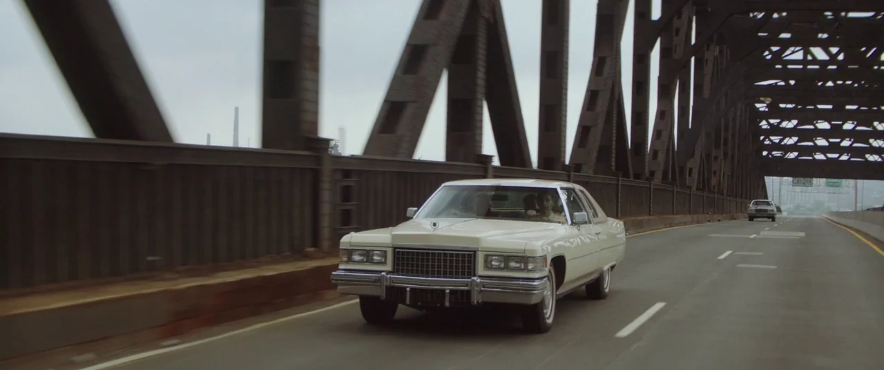God’s Pocket (2014) movie still: wide — A cream-colored 1970s Cadillac Sedan de Ville drives across a large industrial steel bridge. The sky is…