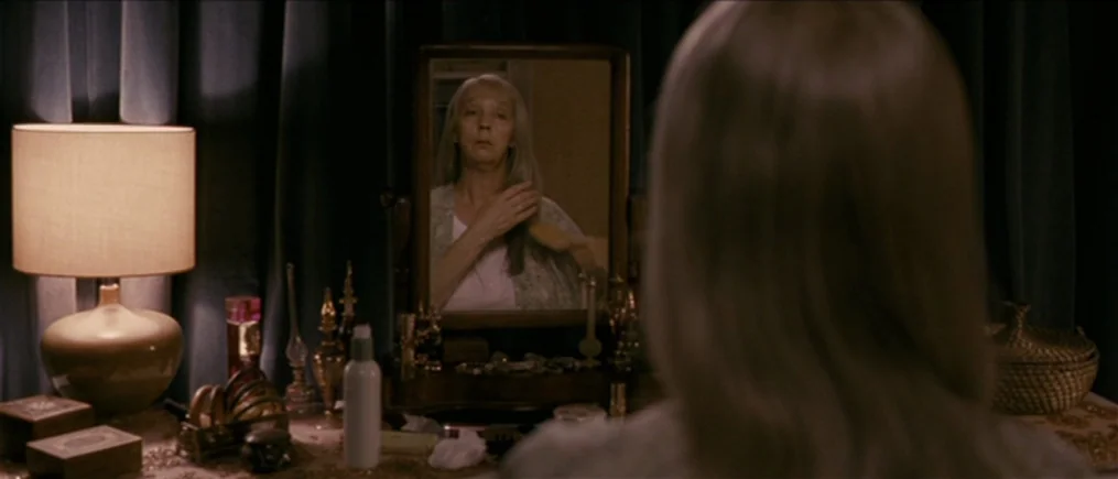 Another Year (2010) movie still: medium — A dimly lit vanity area with dark blue curtains and a table lamp. An older woman brushes her long gray…