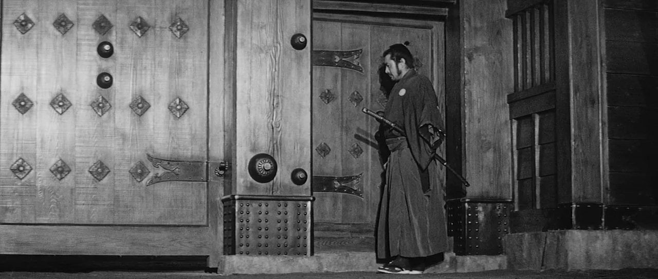 Sanjuro (1962) movie still: fullbody — A man stands before a massive, reinforced wooden fortress gate with decorative metal studs. He pauses near…