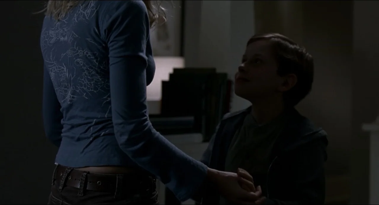 The Ring Two (2005) movie still: medium — A dimly lit indoor space with dark wall shadows and minimal furniture visible. A blonde woman and a young…