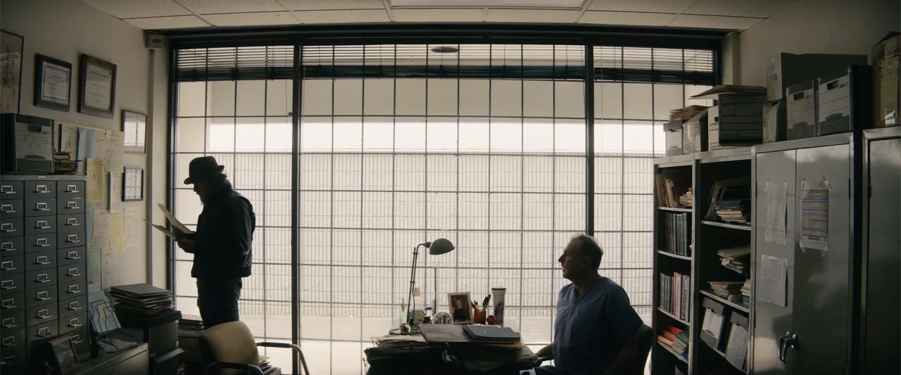 Last Looks (2022) movie still: wide — A cluttered small office contains a metal desk, filing cabinets, and a rear window wall. A man in a hat stands…
