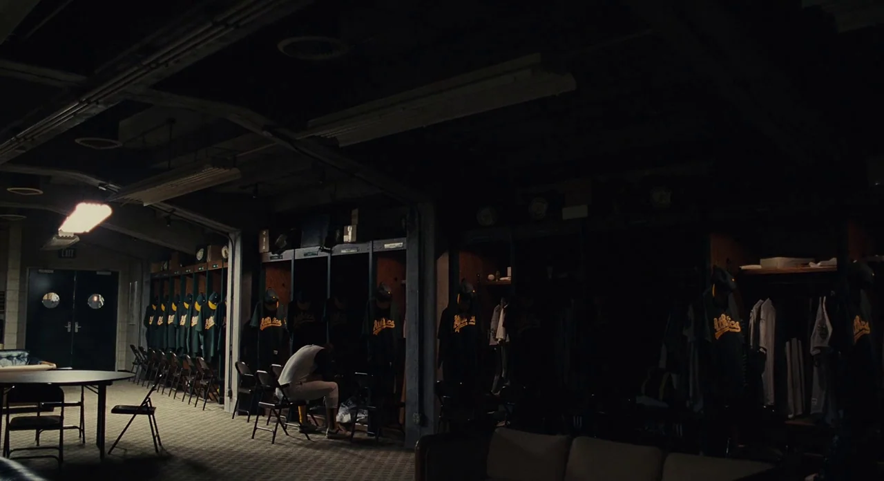Moneyball (2011) movie still: wide — A dimly lit professional sports team dressing room with rows of dark wooden lockers. A single baseball player…