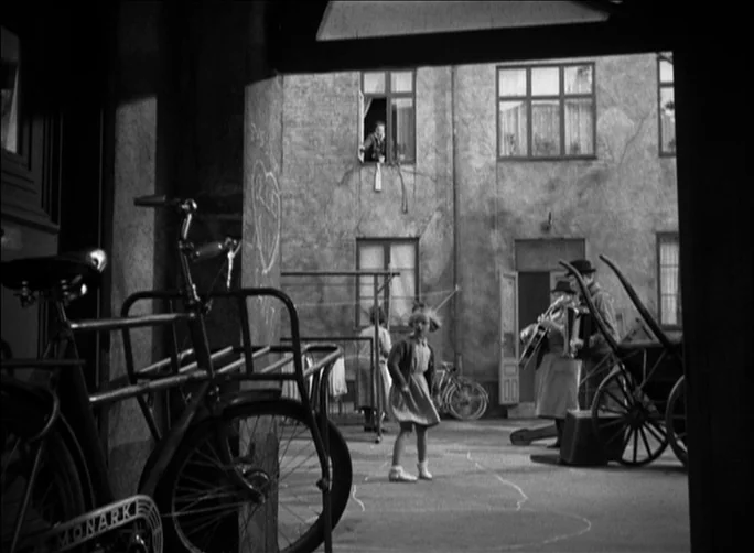 Summer with Monika (1953) movie still: wide — A stone-walled apartment courtyard features a child playing near a chalk-drawn game board. A man with…