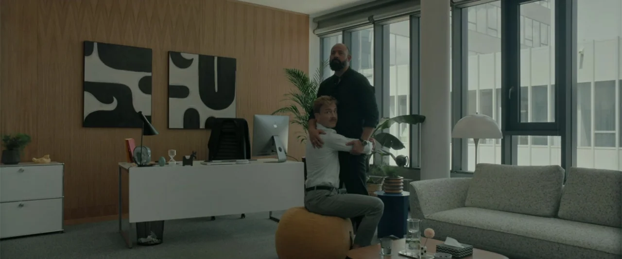 Peacock (2010) movie still: medium — A modern, minimalist office space with wood-paneled walls and large windows overlooking an exterior. Two men…