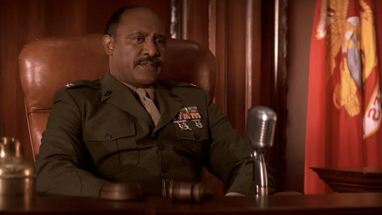 A Few Good Men (1992) movie still: medium — A dark wood-paneled office contains a large leather chair and a military flag. A man in a green military…