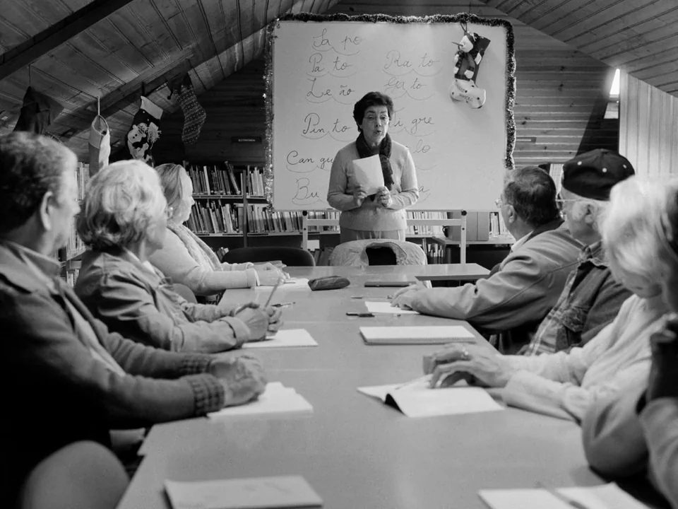 Tabu (2012) movie still: medium — A group of older adults sit around a long table in an attic-like room. A woman stands at the head of the table…