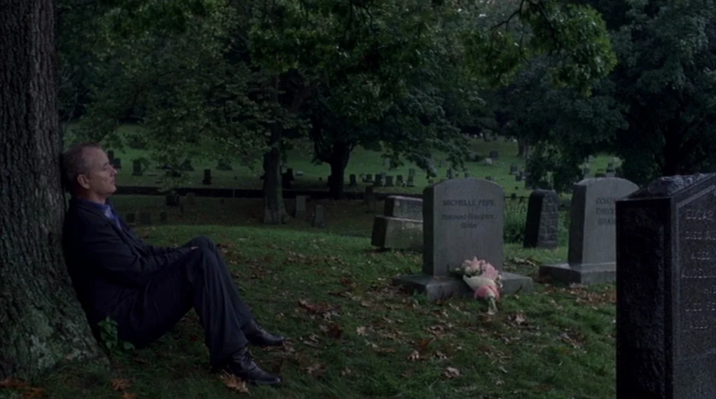 Broken Flowers movie still: wide — A grassy cemetery area is filled with numerous grey headstones under large leafy trees. An older man in a suit…
