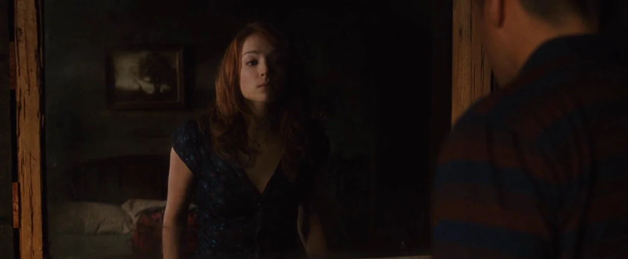 The Cabin In The Woods (2012) movie still: medium — A dimly lit room with a mirror reflecting a woman and an obscured man. The woman stares at her…