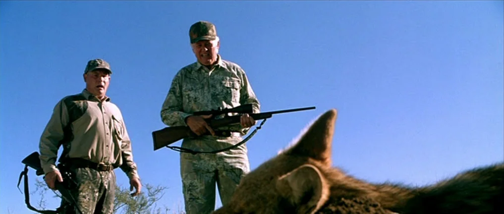 The Three Burials of Melquiades Estrada (2005) movie still: medium — Two men in camouflage gear stand outdoors under a bright blue sky. They hold…