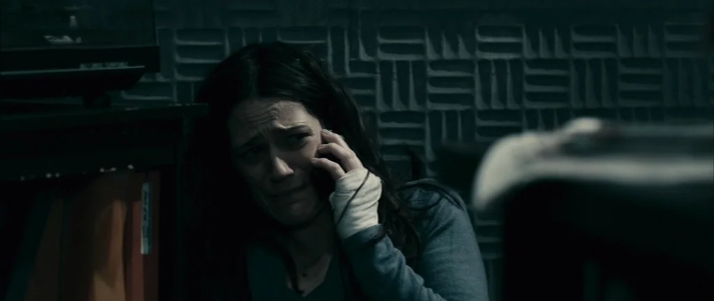 Pontypool (2008) movie still: medium — A dimly lit space features a textured wall with a repetitive geometric pattern. A woman with a bandaged hand…