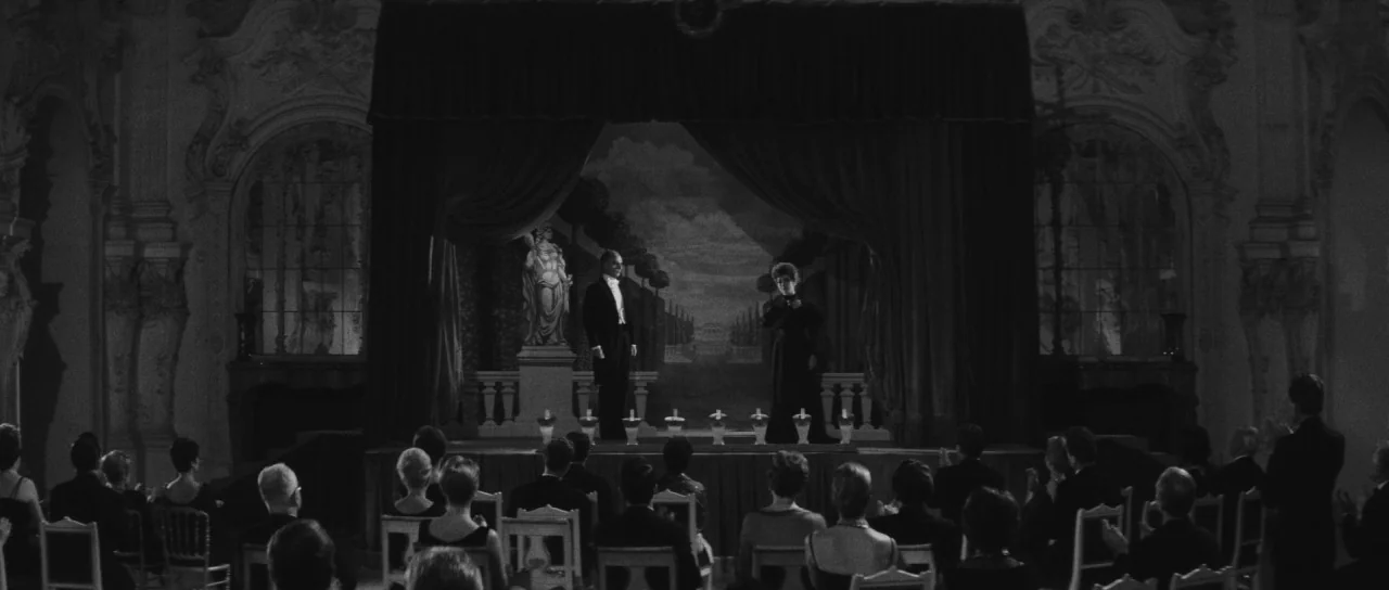 Last Year at Marienbad (1961) movie still: wide — A dark, ornate ballroom features a small wooden stage with heavy curtains and statuary. A man in a…